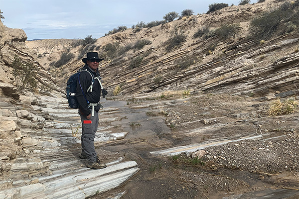 A man stands among obviously striated rock formations in an arid environment.