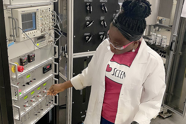 A woman wearing a lab coat adjusts dials on electronic equipment.