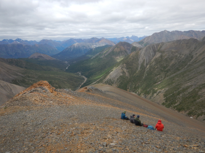 Four people in outdoor clothing sit, backs facing the camera, on a steep slope in the mountains. The ridge they’re on is made of dark gray and orange rocks. In the distance, rugged gray peaks stand above dark green valleys and trees. It’s an overcast day; it looks chilly.