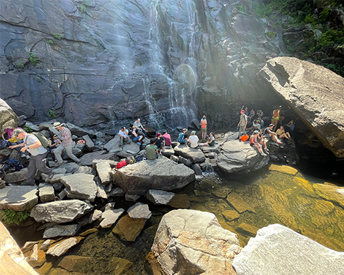A group of about twenty people rest and converse by a small waterfall and rocky natural pool.