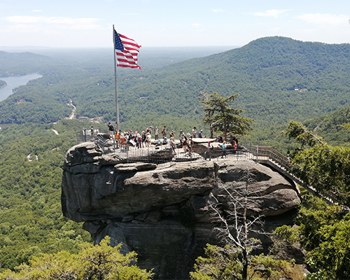 A group of about twenty people explores a large rocky outcrop protruding above a dense forest.
