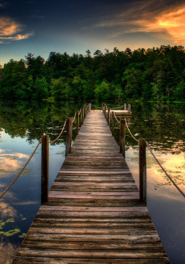 A gently aged dock extends into the still waters of a lushly forested lake at sunset.