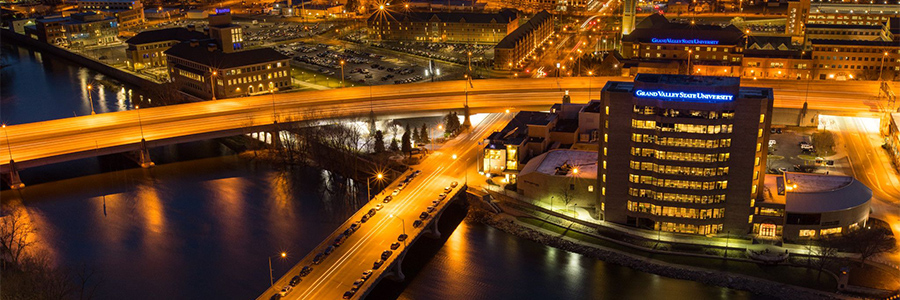 The orange glow of streetlights illuminates two bridges over water in an urban area, and a building sits on the banks with blue signage reading Grand Valley State University.