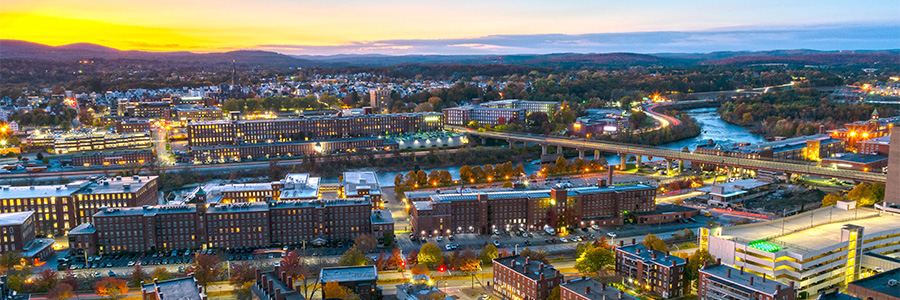 A river snakes through a small urban downtown filled with red brick buildings and glowing lights at sunset.