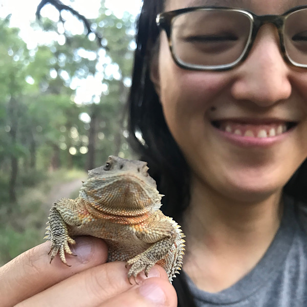 A young woman wearing glasses holds up a horned lizard toward the camera and smiles.