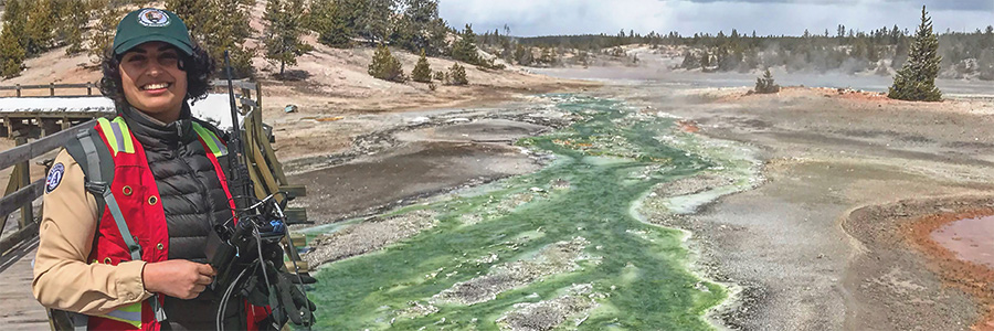 A woman in a National Park Service uniform stands with scientific gear in front of geothermal features.