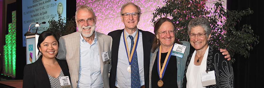 Five GSA Members, two with medals, stand together with a conference stage and lecturn behind them.
