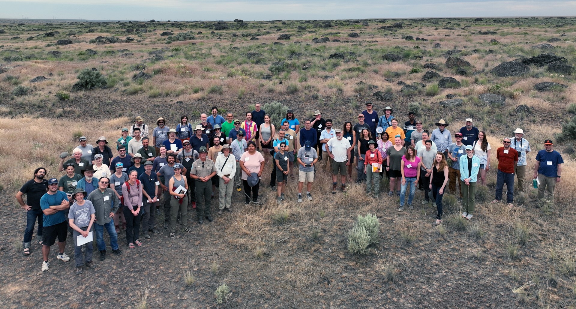 A group of about 60 people smile up at an elevated camera, presumably a drone, with a semi-arid sage steppe in the background dotted with large, dark, rock outcrops.