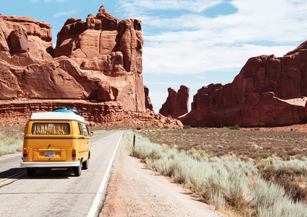 A yellow van with a white roof and a blue surfboard on top drives along a paved road through a desert landscape. Tall, reddish-brown rock formations rise on either side of the road under a partly cloudy sky, evoking a sense of adventure and travel through a scenic, arid region.