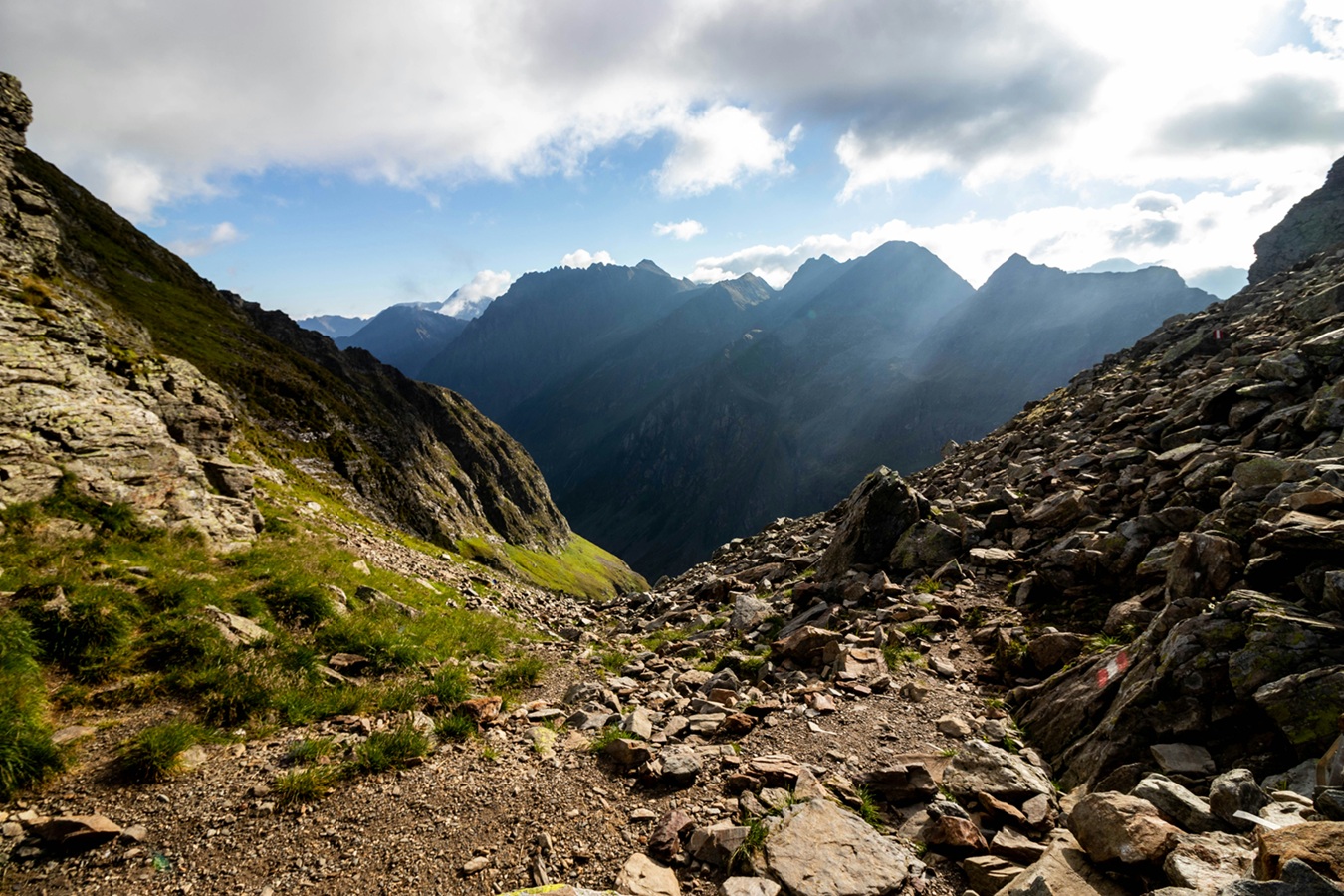 In the left foreground is a green hillside; on the right is a rocky hillside with slices of rock coming loose and sliding down. In the background is a mountain range with blue sky and clouds above.