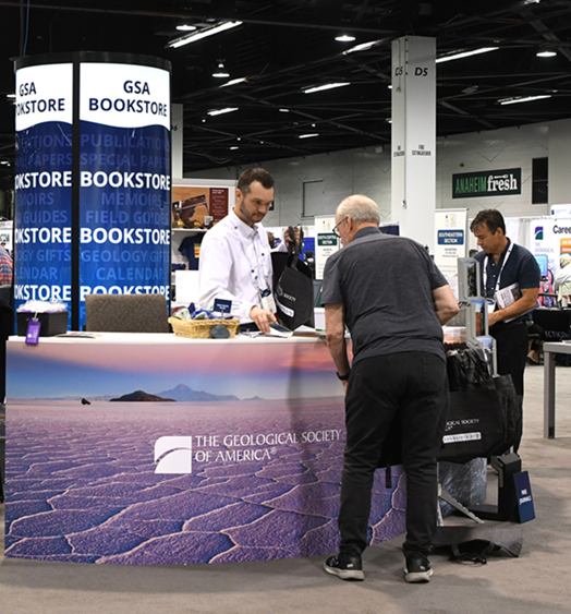 GSA Bookstore at Connects 2024. Attendees are interacting with GSA staff at the booth, which displays colorful geological imagery, promotional materials, and a prominent GSA logo.