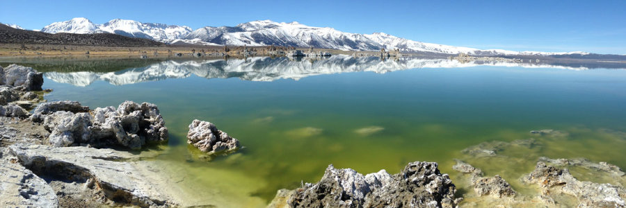 Mineral-encrusted rocks in the foreground of a still lake with snowy mountains against the horizon.