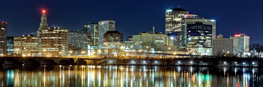 A colorful, brightly lit city skyline glows against a twilit sky and is reflected in a body of water.