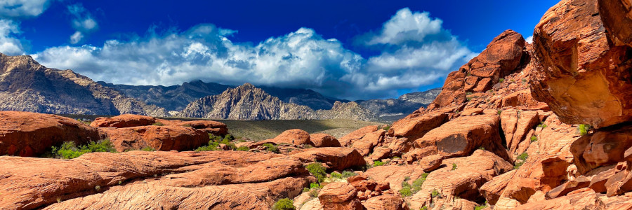 Red sandstone features dominate the foreground of a landscape with rugged mountains under a blue, partly cloudy sky.