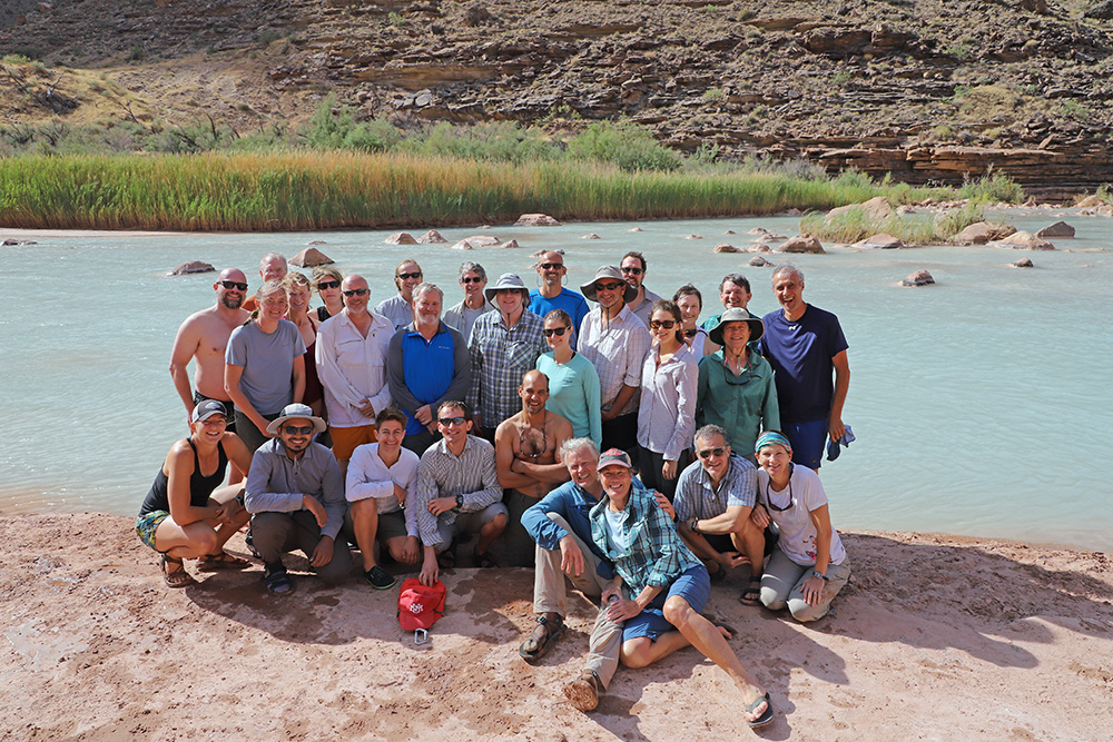 A group of about 30 people smile on the sandy banks of a river.