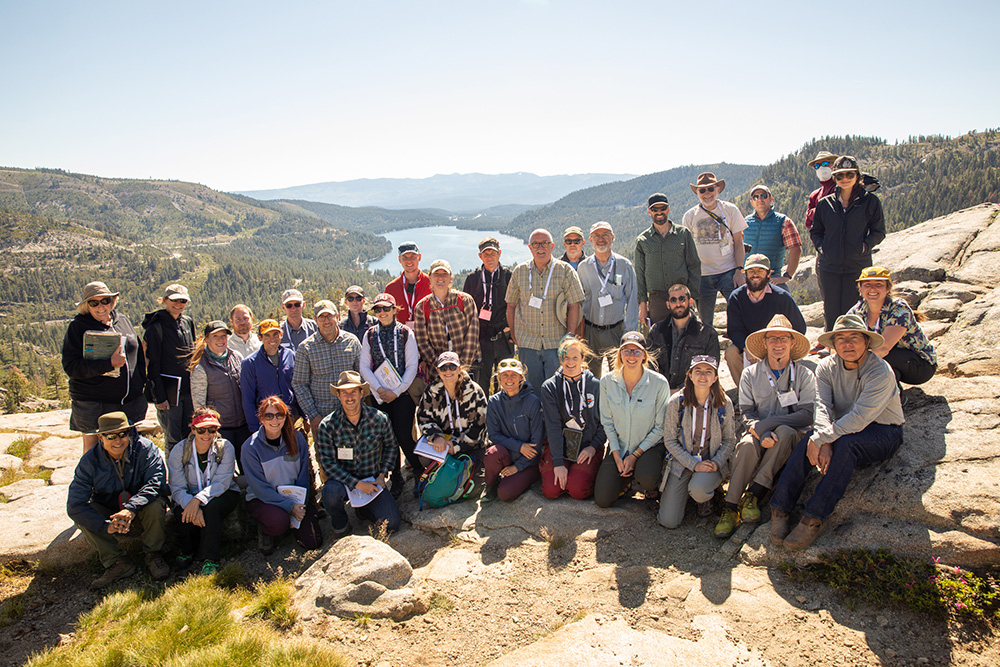 A group of about 35 people smile on a rocky outcrop overlooking a pine forest and mountain lake.